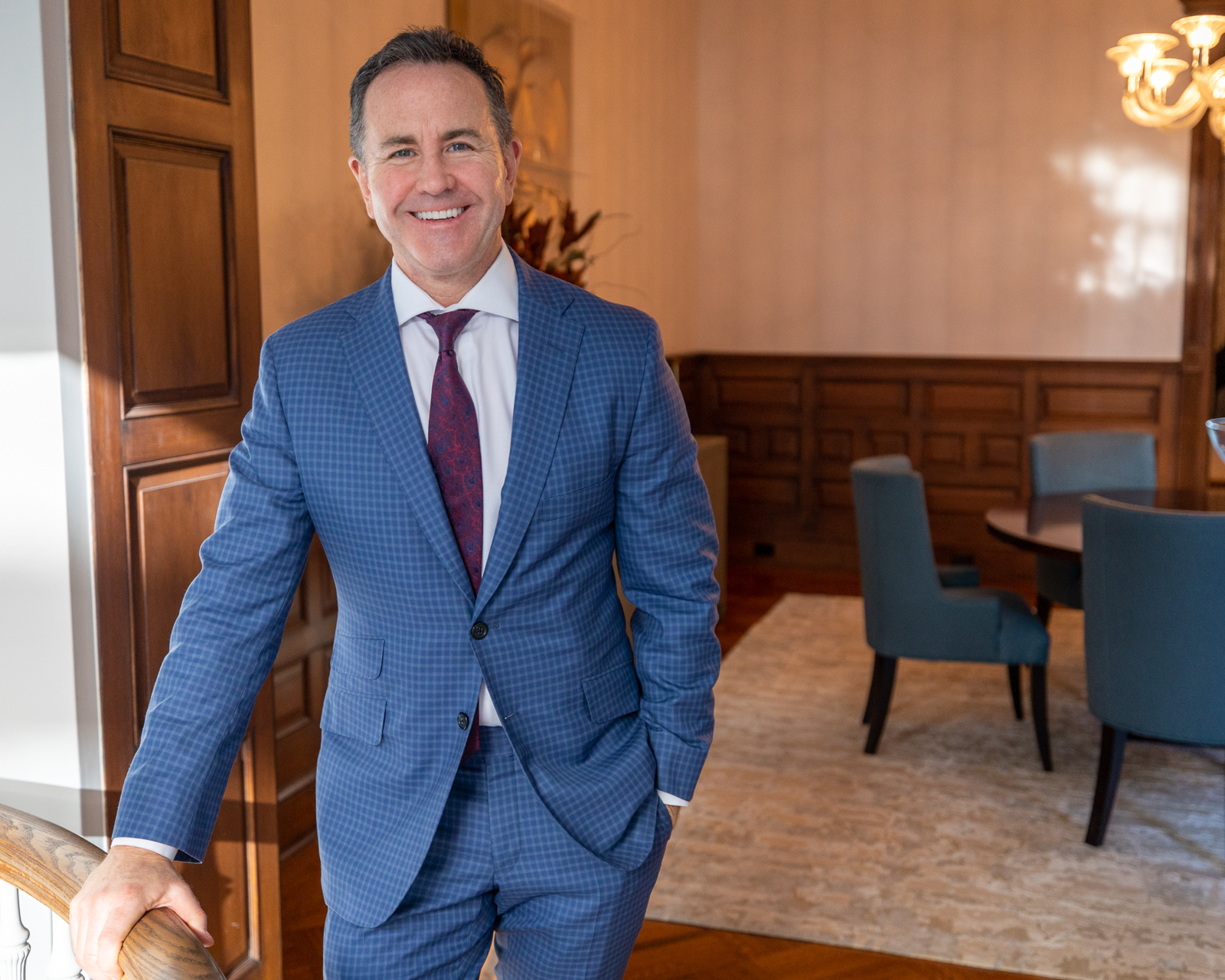 A smiling man in a blue checked suit rests a hand on a wooden stair railing, with a dining room and teal chairs behind him.